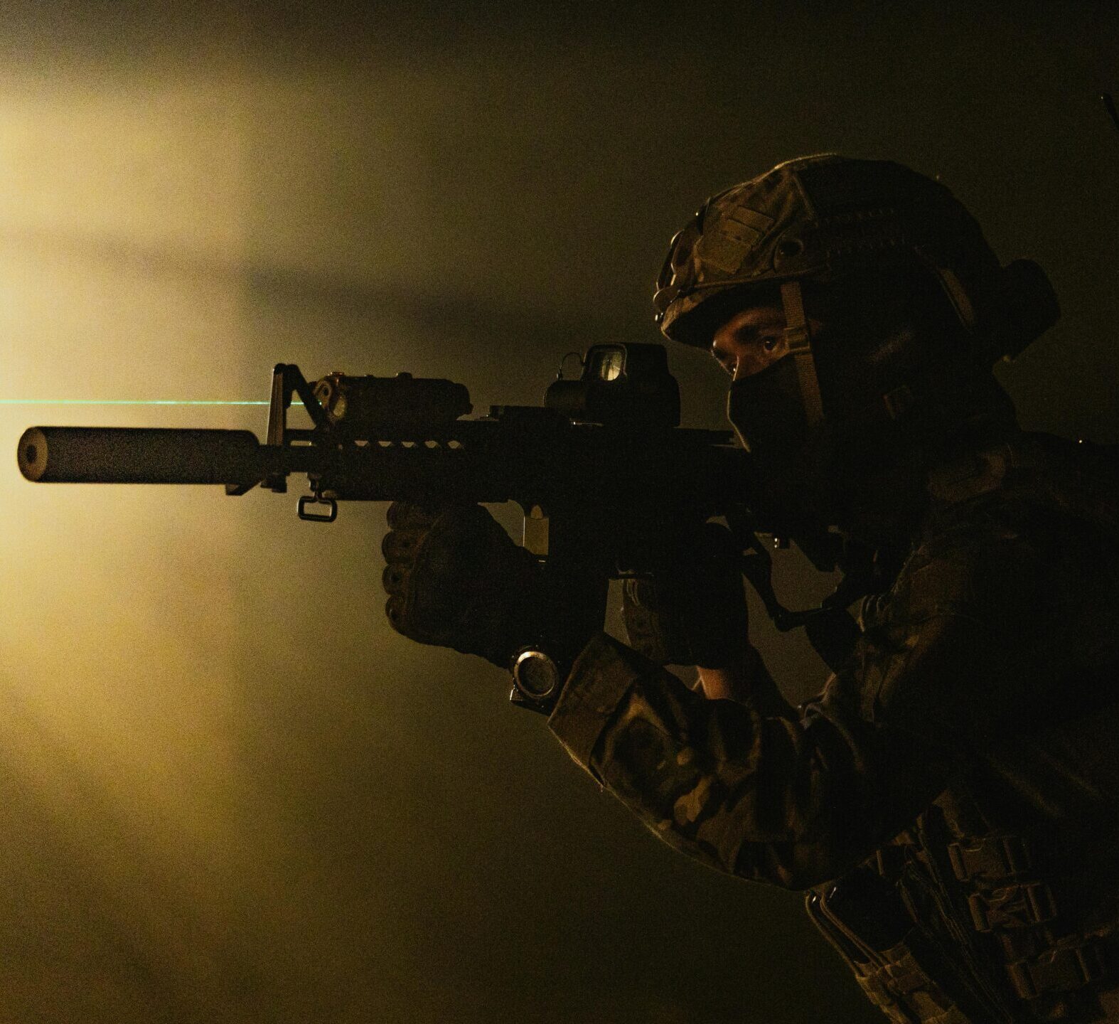 A soldier in tactical gear aims a weapon with a laser sight in low light conditions.